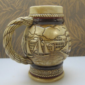 May include: A ceramic beer stein with a rope-like handle and a detailed scene of sailboats and a town on the front. The stein is decorated with brown and red accents and the text "Old Headded Ketch" is visible on the bottom.