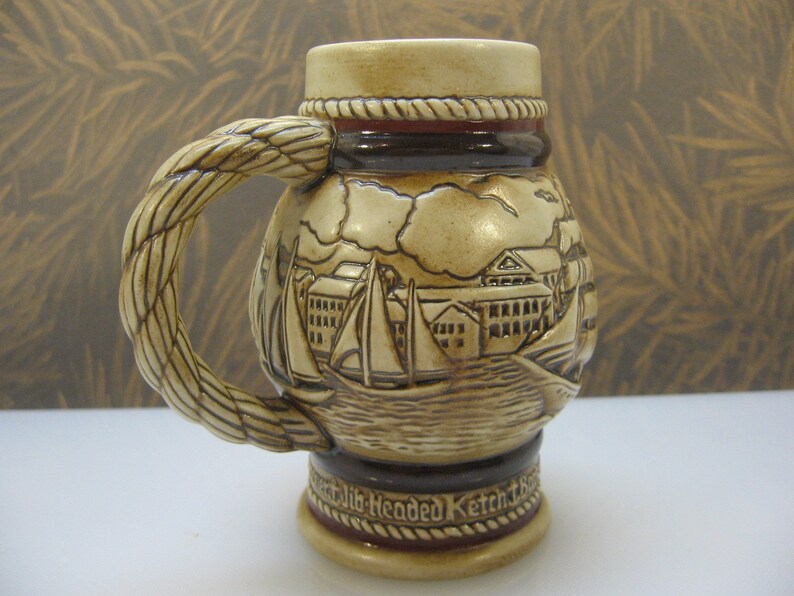 May include: A ceramic beer stein with a rope-like handle and a detailed scene of sailboats and a town on the front. The stein is decorated with brown and red accents and the text "Old Headded Ketch" is visible on the bottom.