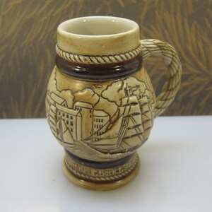May include: A ceramic beer stein with a brown and beige color scheme. The stein features a rope-like handle and a detailed illustration of a ship and buildings. The text "Sailing C. Bark T. Schooner" is visible at the bottom.