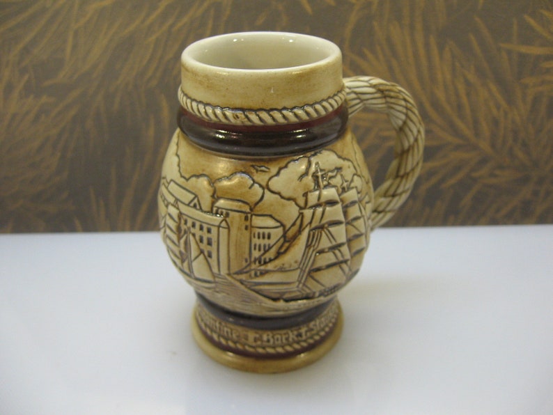 May include: A ceramic beer stein with a brown and beige color scheme. The stein features a rope-like handle and a detailed illustration of a ship and buildings. The text "Sailing C. Bark T. Schooner" is visible at the bottom.