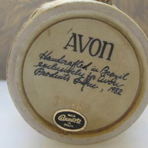 May include: A beige ceramic mug with a brown handle. The bottom of the mug has a sticker that says "Made in Brazil" and the text "Avon Handcrafted in Brazil exclusively for Avon Products, 1982".
