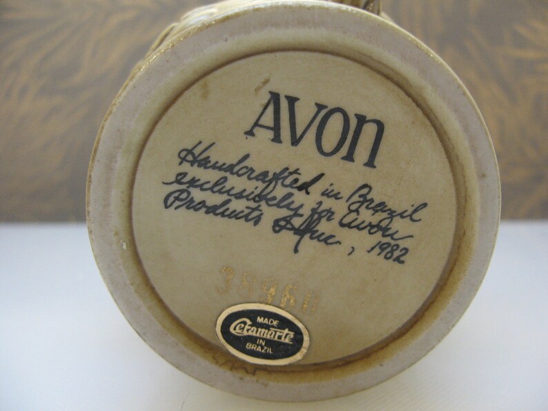 May include: A beige ceramic mug with a brown handle. The bottom of the mug has a sticker that says "Made in Brazil" and the text "Avon Handcrafted in Brazil exclusively for Avon Products, 1982".
