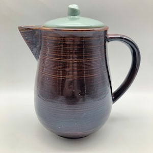 May include: A brown ceramic pitcher with a light green lid. The pitcher has a wide base and a narrow neck. The lid has a small knob on top.