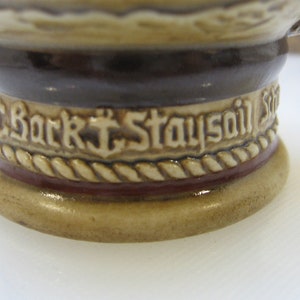 May include: Close-up of a ceramic stein with a brown and white rope design and the text "Bark Staysail" in white.