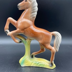 May include: A glossy ceramic horse figurine in a rearing pose, featuring a chestnut brown body with a white mane and tail. The horse stands on a base painted with yellow and green accents, with dark gray detailing on the hooves and muzzle.