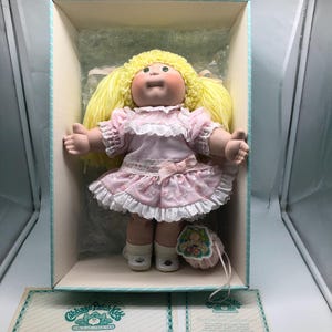 May include: A Cabbage Patch Kid doll with blonde hair, green eyes, and a pink dress. The doll is in a box with a certificate of authenticity. The certificate is signed by Xavier Roberts.