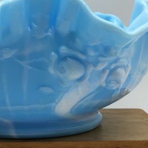 May include: A light blue glass bowl with a ruffled edge and white floral design. The bowl is made of opalescent glass and has a vintage look.