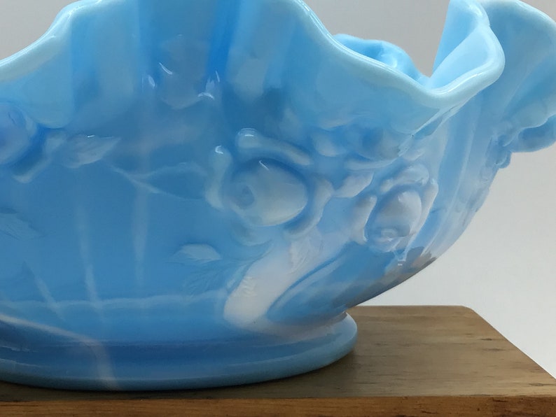May include: A light blue glass bowl with a ruffled edge and white floral design. The bowl is made of opalescent glass and has a vintage look.
