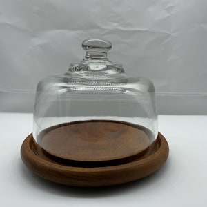 May include: A clear glass cloche with a rounded top and a small knob sits atop a round, dark wooden base. The base has a raised edge. The cloche is designed to cover and protect food items.