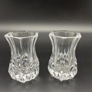 May include: Two clear glass vessels featuring a distinct hourglass shape with a wide, hexagonal rim and a tapered waist. The lower portion of each glass is decorated with a faceted, diamond-cut pattern that catches the light, creating a textured appearance.