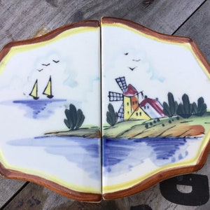 May include: Two ceramic tiles with a hand-painted design. The left tile features two sailboats on a blue body of water with a green island in the foreground. The right tile features a windmill and a small village on a green hill with a blue body of water in the foreground. Both tiles have a brown border.
