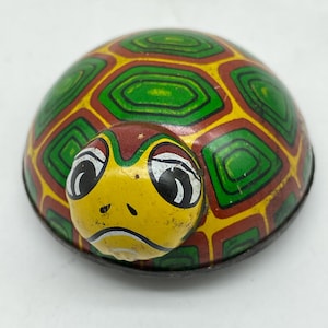 May include: A vintage wind-up toy turtle. The turtle's shell is green, yellow, and brown with a geometric pattern. The turtle's head is yellow with black eyes and a sad expression. The toy is made of metal.