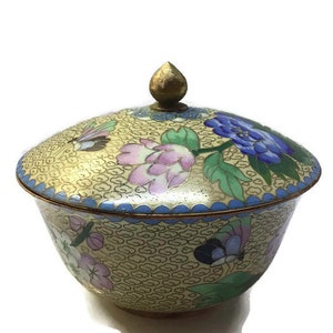 May include: A small, round, gold-colored porcelain bowl with a lid. The bowl and lid are decorated with a floral pattern in shades of blue, pink, and green. The bowl has a scalloped edge.