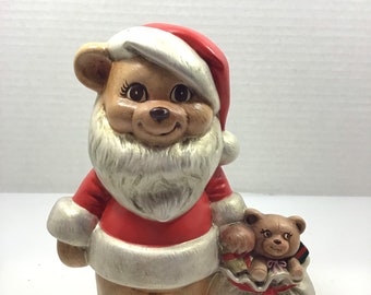 May include: A ceramic bear figurine dressed as Santa Claus, wearing a red and white suit and hat. The bear has a white beard and is holding a sack with a smaller teddy bear inside. The figurine is a festive holiday decoration.