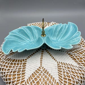 May include: A turquoise ceramic leaf-shaped serving dish with a gold-colored handle. The dish is placed on a wooden surface covered with a white crocheted doily. The dish has detailed leaf vein patterns.