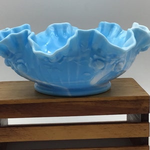 May include: A light blue glass bowl with a ruffled edge and a white floral design. The bowl is sitting on a wooden stand.