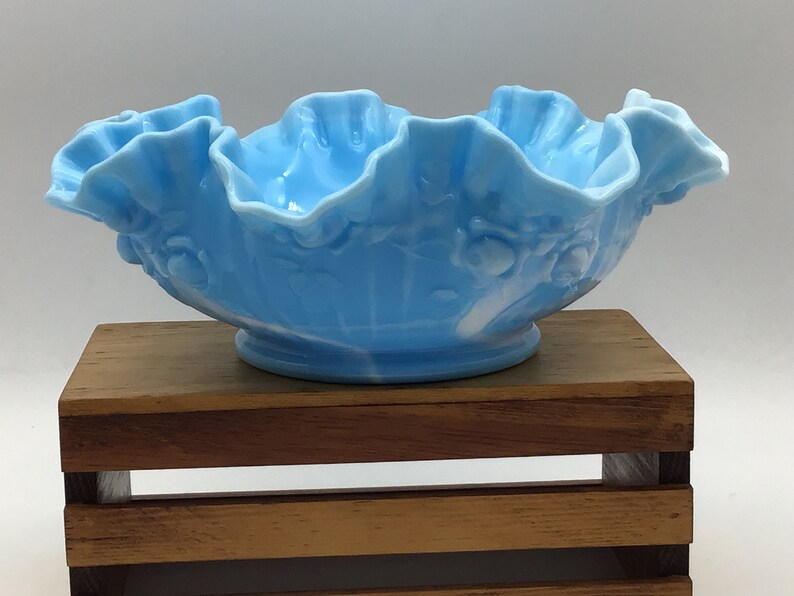 May include: A light blue glass bowl with a ruffled edge and a white floral design. The bowl is sitting on a wooden stand.