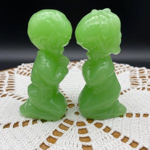 May include: Two translucent light green glass figurines depicting figures in a kneeling, praying position. The figures are smooth, molded glass with rounded features, positioned facing each other, showcasing a consistent bright lime green color throughout.