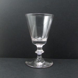 May include: A clear, stemmed glass with a flared bowl and a detailed stem. The glass sits on a round base. The glass is empty and appears to be made of crystal or a similar transparent material. The background is a solid black.