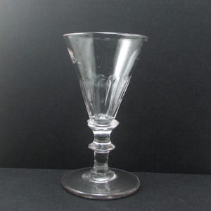 May include: A clear, vintage-style glass with a flared bowl and a detailed stem. The glass features vertical etched lines on the bowl and a round base. The glass is designed for serving beverages.