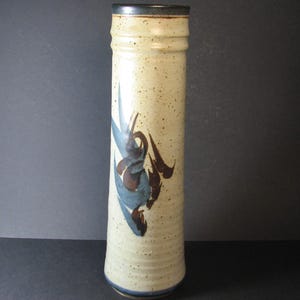May include: A tall, cylindrical ceramic vase with a speckled beige glaze. It features a stylised floral design in shades of blue and brown.
