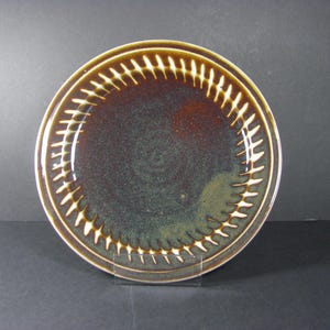 May include: A ceramic plate with a dark brown center and a lighter brown rim. The rim features a repeating pattern of white, stylized leaf shapes. The plate is approximately 10 inches in diameter.