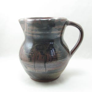 May include: A handmade ceramic pitcher with a dark brown and reddish-brown glaze. The pitcher has a rounded body, a spout, and a curved handle. The surface has a textured appearance with horizontal lines.