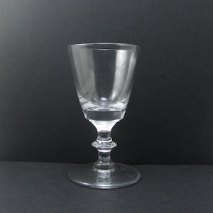 May include: A clear, stemmed glass with a wide, slightly flared bowl. The glass has a short stem with a decorative bulbous design and a circular base. The glass is empty and sits against a dark background.