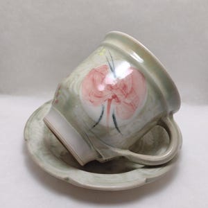 May include: A ceramic teacup and saucer set. The cup and saucer are a pale green with a floral design in pink and black. The cup has a handle and is resting on the saucer.