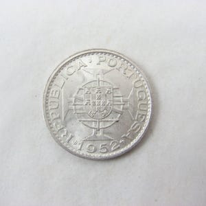 May include: A silver coin from 1952 with the words "REPUBLICA PORTUGUESA" around the edge. The coin features a coat of arms in the center and a cross design. The coin is round and has a textured edge.