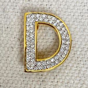 May include: A gold-toned letter "D" brooch, encrusted with clear rhinestones. The brooch has a polished gold border and is set against a neutral textured background. The letter is a classic, elegant design.