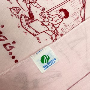 May include: Pink fabric with a red illustration of people camping. The fabric has a tag that reads "GIRL SCOUTS&reg; MADE IN U.S.A." The illustration depicts a camping scene with a tent and cooking equipment.