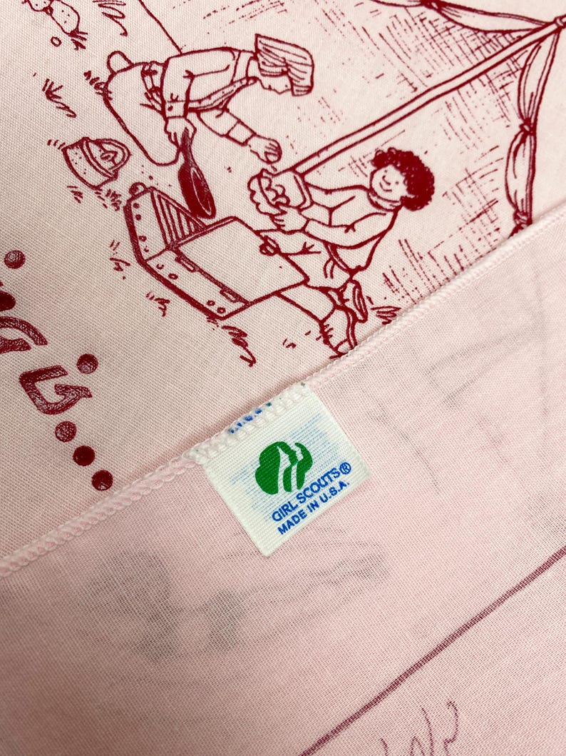 May include: Pink fabric with a red illustration of people camping. The fabric has a tag that reads "GIRL SCOUTS&reg; MADE IN U.S.A." The illustration depicts a camping scene with a tent and cooking equipment.