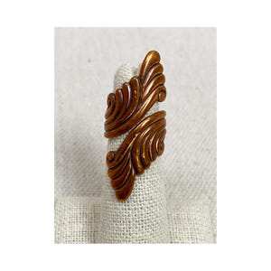 May include: A copper-colored ring with an ornate, swirling leaf design. The ring wraps around the finger, with the leaf-like elements overlapping. The ring is displayed on a neutral-toned fabric.