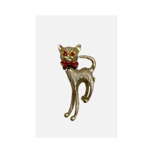 May include: Gold-toned cat brooch with red rhinestone eyes and a red bow tie. The cat has a textured surface and a curved tail. A vintage-style accessory.