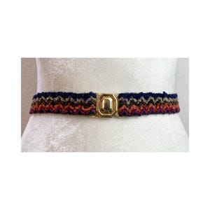May include: A multi-colored crochet belt with a gold-toned rectangular buckle. The belt features a chevron pattern in shades of navy blue, brown, orange, and purple. The belt is displayed on a white background.