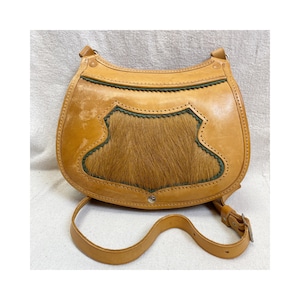May include: Tan leather shoulder bag with a central panel of brown fur. The bag has a curved shape, a long adjustable strap, and green trim. A silver button secures the flap.