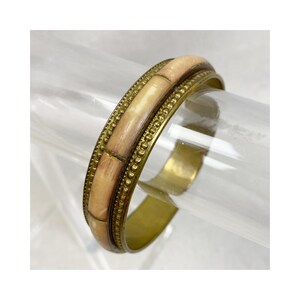 May include: A brass bangle bracelet with a light tan and brown bamboo-style design. The bracelet has a gold-colored metal band with a dotted border. The bamboo-style sections are rectangular and arranged around the bracelet.