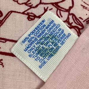 May include: A close-up of a white tag with blue text, showing fabric care instructions. The tag is on a pink fabric with a red design. The tag reads: 50% Polyester/50% Cotton, Machine wash warm, only non-chlorine bleach when needed, tumble dry medium, to iron: place damp cloth on reverse side, use cool setting.