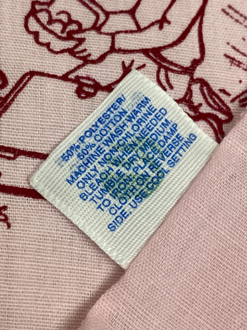 May include: A close-up of a white tag with blue text, showing fabric care instructions. The tag is on a pink fabric with a red design. The tag reads: 50% Polyester/50% Cotton, Machine wash warm, only non-chlorine bleach when needed, tumble dry medium, to iron: place damp cloth on reverse side, use cool setting.