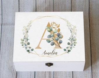 Könnte beinhalten: Weiße Holzkiste mit goldenem geometrischem Rahmen und floralem Design. Der Buchstabe "A" ist in Gold mit einem Aquarell-Blumendesign. Der Name "Amelia" ist in goldener Schrift unter dem Buchstaben geschrieben.