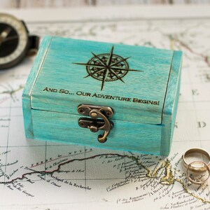 May include: A teal blue wooden box with a compass design and the text "And so... Our Adventure Begins!" engraved on the lid. The box is sitting on a vintage map with two gold wedding bands.