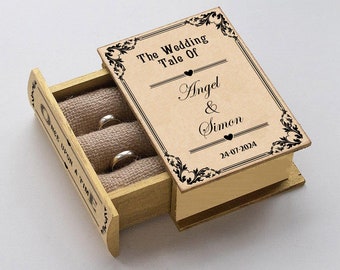 May include: A wooden book-shaped ring box with a drawer that opens to reveal two gold wedding bands. The box is decorated with black floral designs and the text "The Wedding Tale Of Angel & Simon 24-07-2024".