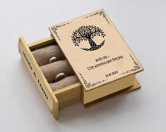 Op de afbeelding: Een houten ringendoos in de vorm van een boek met een Keltisch levensboomontwerp op de omslag. De doos is open en onthult een beige stoffen interieur met twee gouden ringen. De omslag heeft de tekst "And so... the adventure begins" en "22.10.2023".