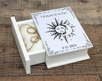 Op de afbeelding: Witte houten ringendoos met een schuifdeksel. Het deksel heeft een zwart-witte illustratie van een zon en maan met de tekst "Till Death Do Us Part" en de datum "11-12-2024".