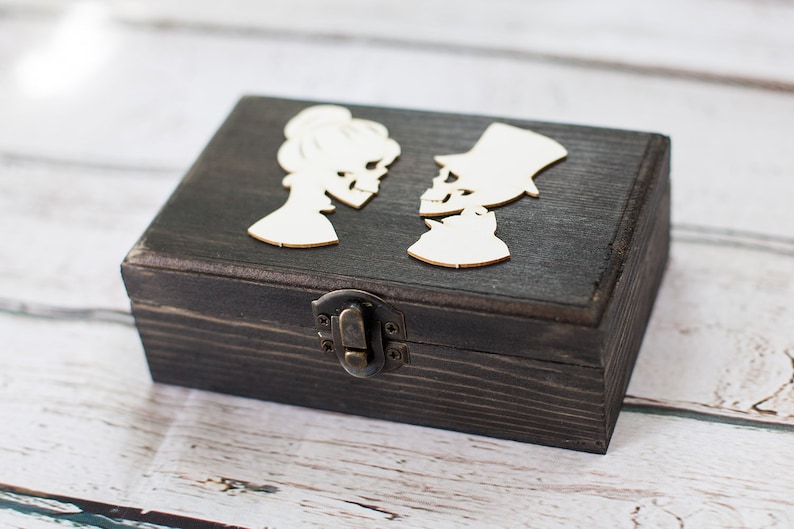 May include: A dark wooden ring box with a hinged lid and a latch closure. The lid features two white skeleton figures facing each other.
