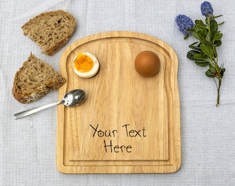 May include: A wooden serving board shaped like a slice of bread with two indentations for eggs. The board has the text "Your Text Here" engraved on it. There are two boiled eggs on the board, one peeled and one unpeeled. There is a spoon and two slices of bread on a white tablecloth.
