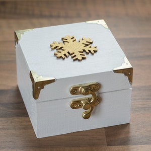 May include: A white wooden box with gold accents and a gold snowflake design on the lid. The box has a gold latch.