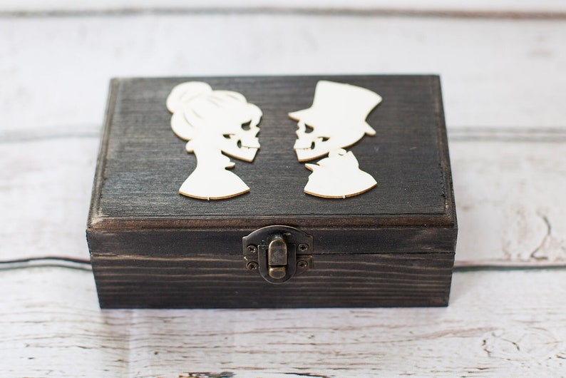 May include: A dark wooden box with a hinged lid and a metal clasp. The box is decorated with two white silhouettes of skeletons, one male and one female, facing each other.