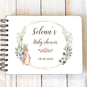 Può includere: Un libro degli ospiti bianco con una cornice geometrica dorata e un'illustrazione ad acquerello di un coniglio e foglie di eucalipto. Il testo "Selena's Baby Shower" e la data "18-09-2023" sono stampati sulla copertina.